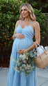 A smiling pregnant woman wearing a blu e tiered halter-neck maternity midi dress, standing in a sunlit garden. The dress features a smocked bust and flowy skirt, ideal for a summer baby shower or outdoor photoshoot.