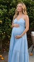 A smiling pregnant woman wearing a blu e tiered halter-neck maternity midi dress, standing in a sunlit garden. The dress features a smocked bust and flowy skirt, ideal for a summer baby shower or outdoor photoshoot.