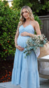 A smiling pregnant woman wearing a blu e tiered halter-neck maternity midi dress, standing in a sunlit garden. The dress features a smocked bust and flowy skirt, ideal for a summer baby shower or outdoor photoshoot.