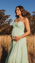 A smiling pregnant woman wearing a green tiered halter-neck maternity midi dress. The dress features a smocked bust and flowy skirt, ideal for a summer baby shower or outdoor photoshoot.
