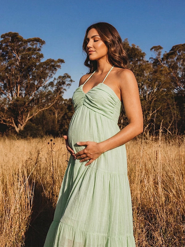 A smiling pregnant woman wearing a green tiered halter-neck maternity midi dress. The dress features a smocked bust and flowy skirt, ideal for a summer baby shower or outdoor photoshoot.