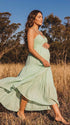 A smiling pregnant woman wearing a green tiered halter-neck maternity midi dress. The dress features a smocked bust and flowy skirt, ideal for a summer baby shower or outdoor photoshoot.