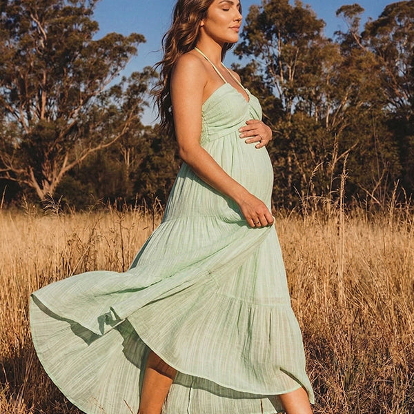 A smiling pregnant woman wearing a green tiered halter-neck maternity midi dress. The dress features a smocked bust and flowy skirt, ideal for a summer baby shower or outdoor photoshoot.
