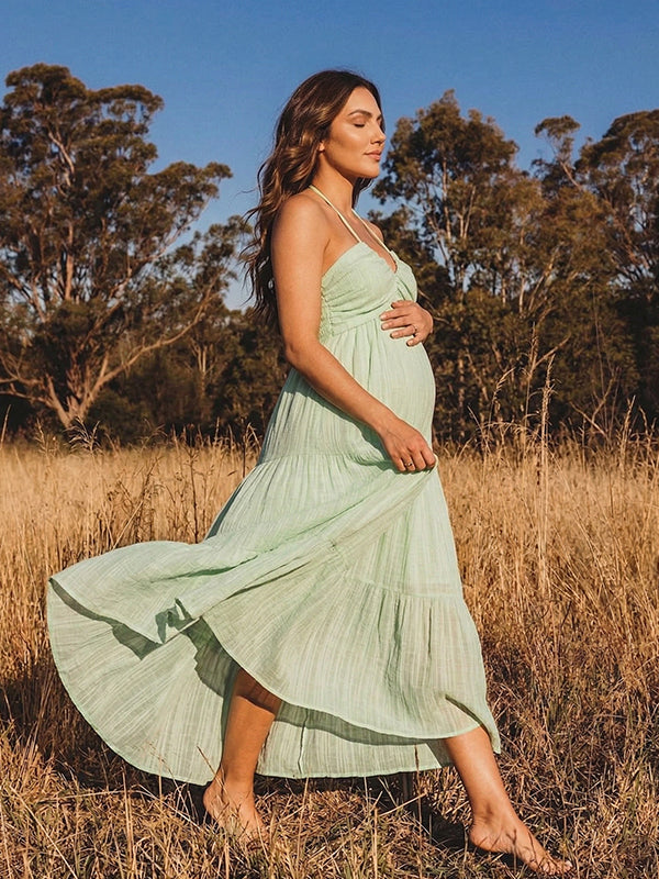 A smiling pregnant woman wearing a green tiered halter-neck maternity midi dress. The dress features a smocked bust and flowy skirt, ideal for a summer baby shower or outdoor photoshoot.