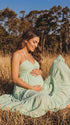 A smiling pregnant woman wearing a green tiered halter-neck maternity midi dress. The dress features a smocked bust and flowy skirt, ideal for a summer baby shower or outdoor photoshoot.