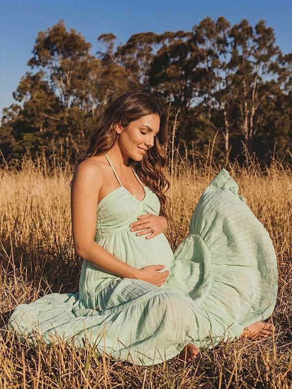 A smiling pregnant woman wearing a green tiered halter-neck maternity midi dress. The dress features a smocked bust and flowy skirt, ideal for a summer baby shower or outdoor photoshoot.