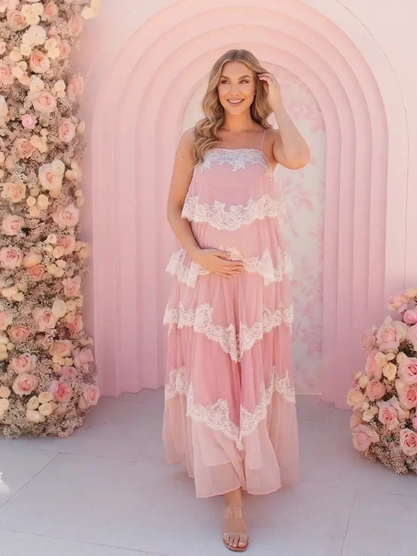 A pink tiered lace maternity maxi dress with layered chiffon, white lace trim, and an adjustable lace-up back for baby shower, maternity photoshoot, gender reveal, and bridal shower.