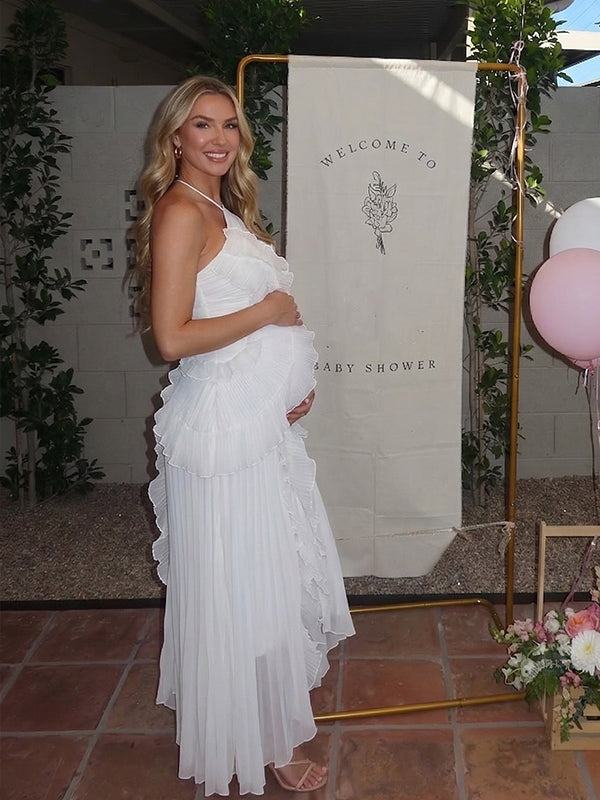 Smiling pregnant woman with wavy blonde hair, wearing an elegant white pleated halter midi dress with a cascading ruffled front and high-low hem, celebrating at a baby shower.