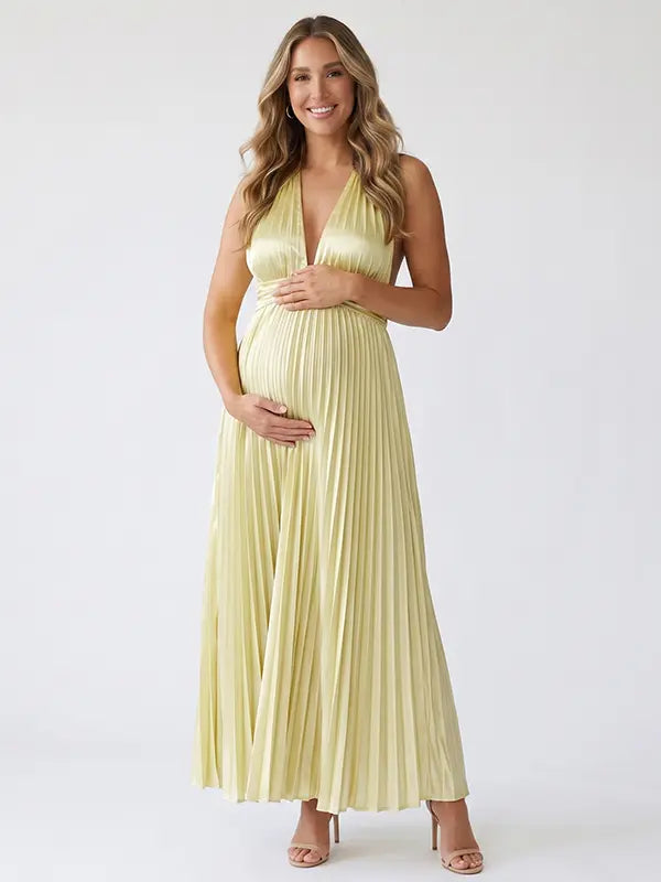 Pregnant model wearing a butter yellow pleated satin halter maternity maxi dress with deep V neckline and flowing skirt for summer baby shower, garden party and maternity photoshoot.