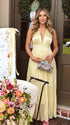 Pregnant model wearing a butter yellow pleated satin halter maternity maxi dress with deep V neckline and flowing skirt for summer baby shower, garden party and maternity photoshoot.
