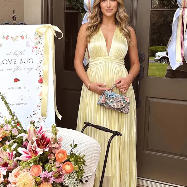 Pregnant model wearing a butter yellow pleated satin halter maternity maxi dress with deep V neckline and flowing skirt for summer baby shower, garden party and maternity photoshoot.