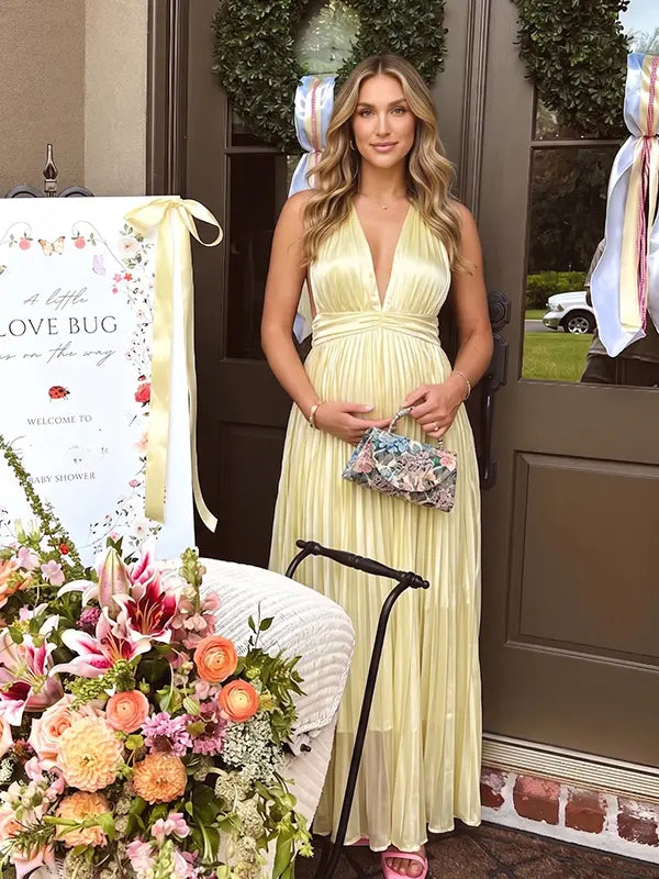 Pregnant model wearing a butter yellow pleated satin halter maternity maxi dress with deep V neckline and flowing skirt for summer baby shower, garden party and maternity photoshoot.