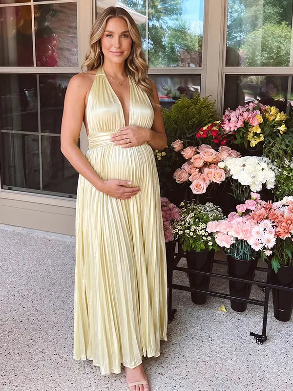 Pregnant model wearing a butter yellow pleated satin halter maternity maxi dress with deep V neckline and flowing skirt for summer baby shower, garden party and maternity photoshoot.