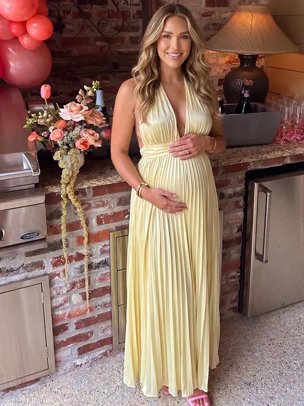 Pregnant model wearing a butter yellow pleated satin halter maternity maxi dress with deep V neckline and flowing skirt for summer baby shower, garden party and maternity photoshoot.