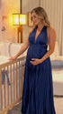 Pregnant model wearing a navy blue pleated satin halter maternity maxi dress with deep V neckline and flowing skirt for wedding guest, maternity photoshoot and formal occasions.