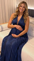 Pregnant model wearing a navy blue pleated satin halter maternity maxi dress with deep V neckline and flowing skirt for wedding guest, maternity photoshoot and formal occasions.