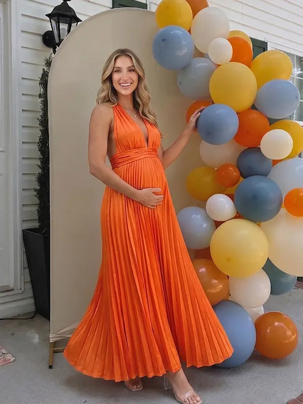 Pregnant model wearing an orange pleated satin halter maternity maxi dress with deep V neckline and flowing skirt for fall baby shower, pumpkin patch photos and maternity photography.