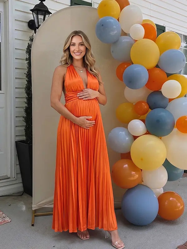 Pregnant model wearing an orange pleated satin halter maternity maxi dress with deep V neckline and flowing skirt for fall baby shower, pumpkin patch photos and maternity photography.