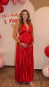 Pregnant model wearing a red pleated satin halter maternity maxi dress with deep V neckline, empire waist and flowing skirt for baby shower, gender reveal and maternity photoshoot.