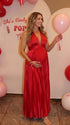 Pregnant model wearing a red pleated satin halter maternity maxi dress with deep V neckline, empire waist and flowing skirt for baby shower, gender reveal and maternity photoshoot.