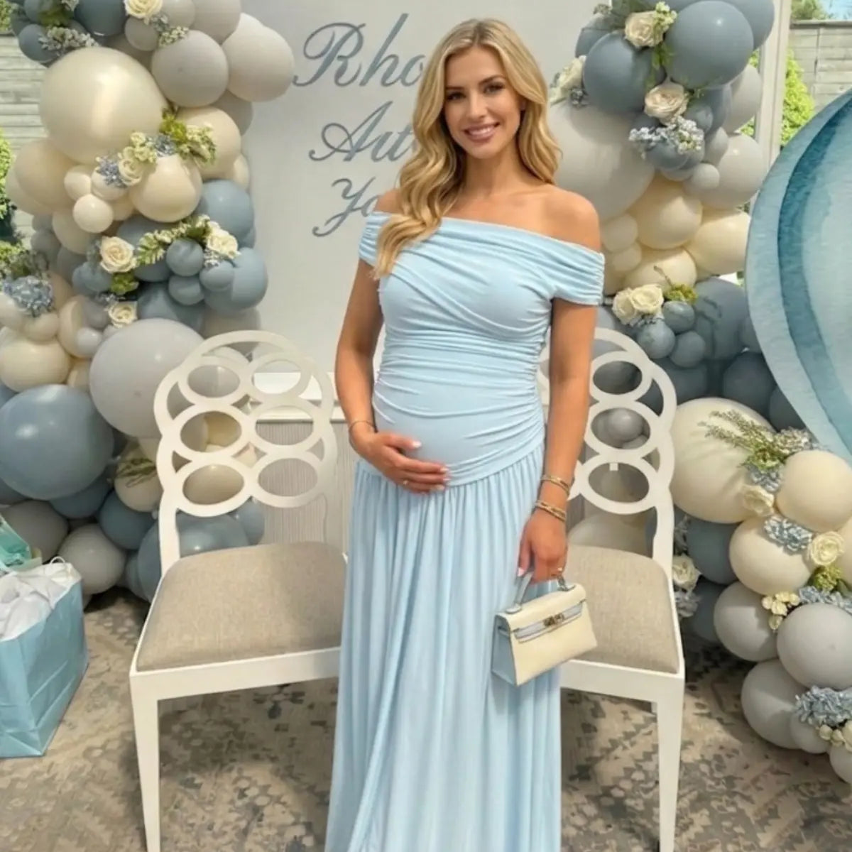 Pregnant woman in a sky blue off-the-shoulder poly-spandex maternity maxi dress with ruched detailing, posing at a baby shower with blue balloon decor.