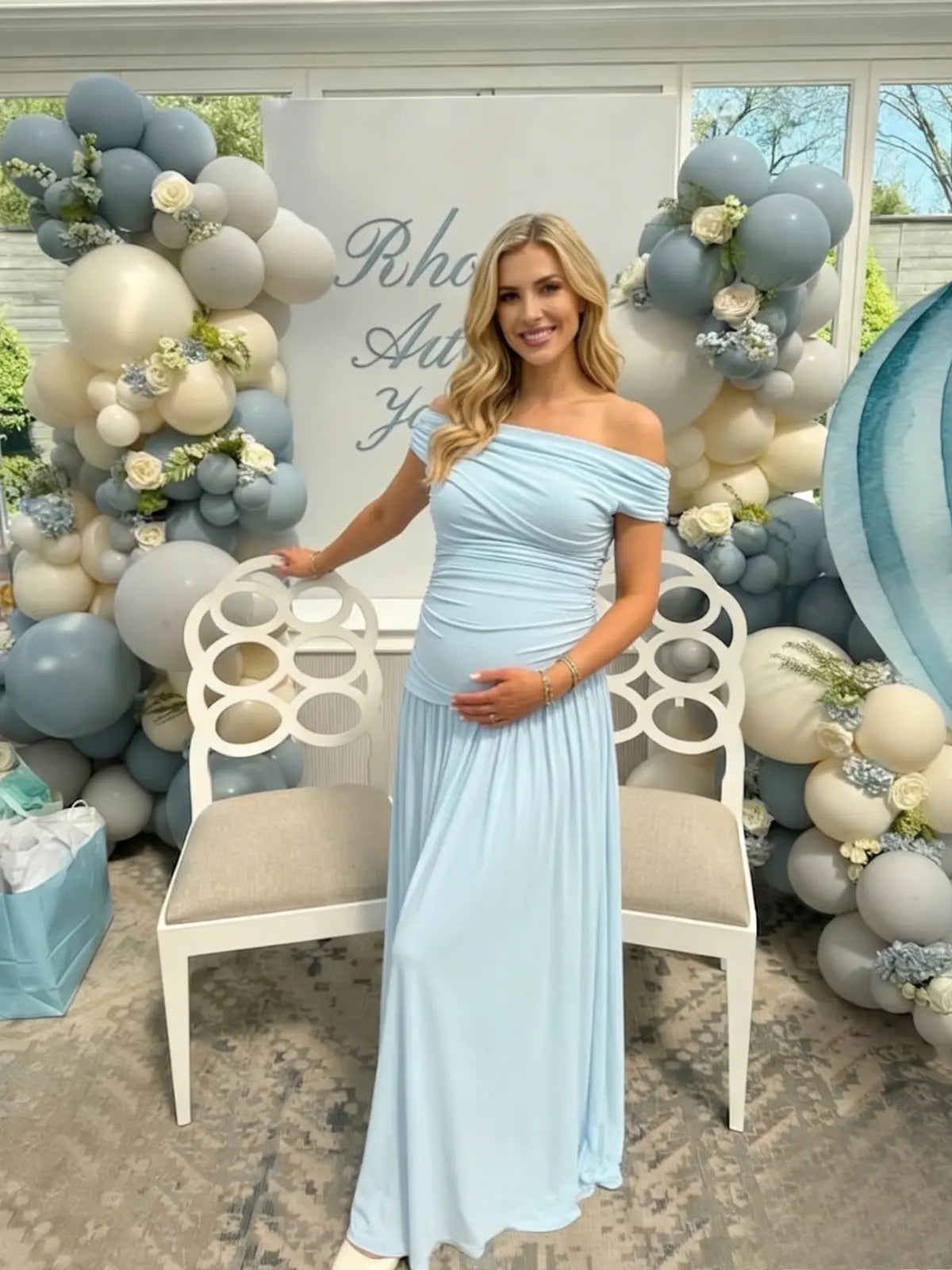Pregnant woman in a sky blue off-the-shoulder poly-spandex maternity maxi dress with ruched detailing, posing at a baby shower with blue balloon decor.