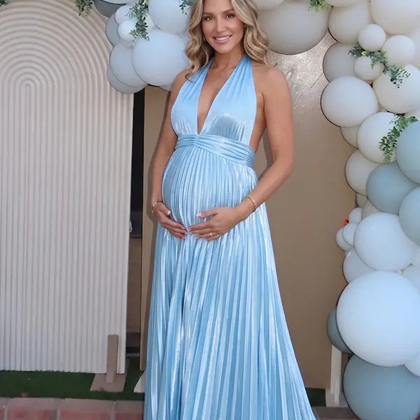 Pregnant model wearing a sky blue pleated satin halter maternity maxi dress with deep V neckline and flowing skirt for beach maternity photoshoot, coastal baby shower and destination pregnancy photography.