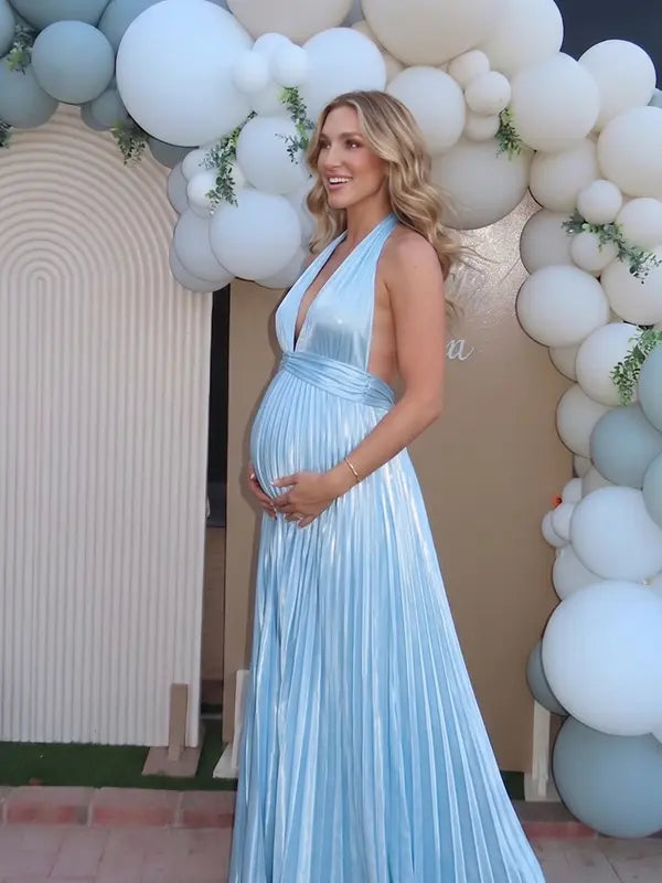 Pregnant model wearing a sky blue pleated satin halter maternity maxi dress with deep V neckline and flowing skirt for beach maternity photoshoot, coastal baby shower and destination pregnancy photography.