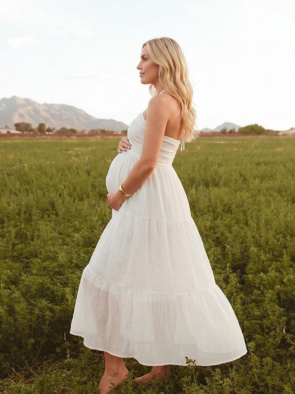 A smiling pregnant woman wearing a yellow tiered halter-neck maternity midi dress. The dress features a smocked bust and flowy skirt, ideal for a summer baby shower or outdoor photoshoot.