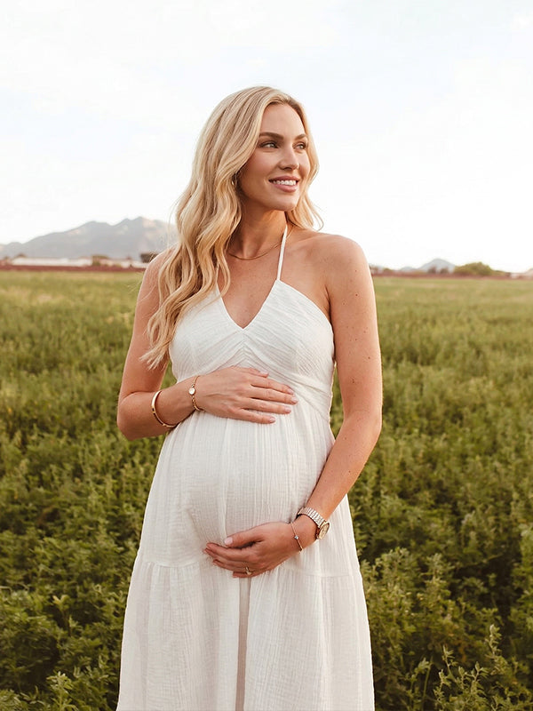 A smiling pregnant woman wearing a yellow tiered halter-neck maternity midi dress. The dress features a smocked bust and flowy skirt, ideal for a summer baby shower or outdoor photoshoot.