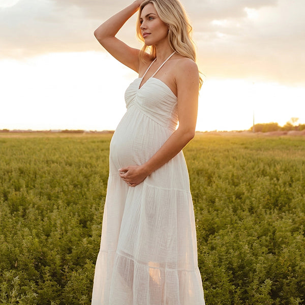 A smiling pregnant woman wearing a yellow tiered halter-neck maternity midi dress. The dress features a smocked bust and flowy skirt, ideal for a summer baby shower or outdoor photoshoot.