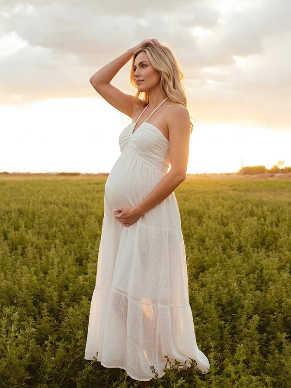 A smiling pregnant woman wearing a yellow tiered halter-neck maternity midi dress. The dress features a smocked bust and flowy skirt, ideal for a summer baby shower or outdoor photoshoot.
