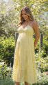 A smiling pregnant woman wearing a bright yellow tiered halter-neck maternity midi dress, standing in a sunlit garden. The dress features a smocked bust and flowy skirt, ideal for a summer baby shower or outdoor photoshoot.