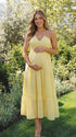 A smiling pregnant woman wearing a bright yellow tiered halter-neck maternity midi dress, standing in a sunlit garden. The dress features a smocked bust and flowy skirt, ideal for a summer baby shower or outdoor photoshoot.