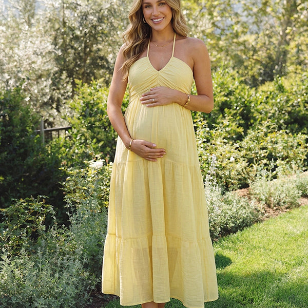 A smiling pregnant woman wearing a bright yellow tiered halter-neck maternity midi dress, standing in a sunlit garden. The dress features a smocked bust and flowy skirt, ideal for a summer baby shower or outdoor photoshoot.