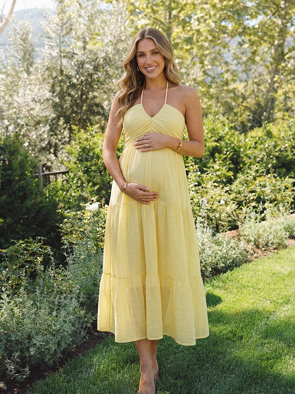 A smiling pregnant woman wearing a bright yellow tiered halter-neck maternity midi dress, standing in a sunlit garden. The dress features a smocked bust and flowy skirt, ideal for a summer baby shower or outdoor photoshoot.