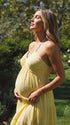 A smiling pregnant woman wearing a bright yellow tiered halter-neck maternity midi dress, standing in a sunlit garden. The dress features a smocked bust and flowy skirt, ideal for a summer baby shower or outdoor photoshoot.