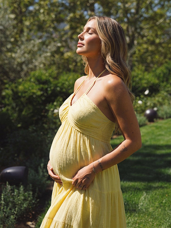 A smiling pregnant woman wearing a bright yellow tiered halter-neck maternity midi dress, standing in a sunlit garden. The dress features a smocked bust and flowy skirt, ideal for a summer baby shower or outdoor photoshoot.
